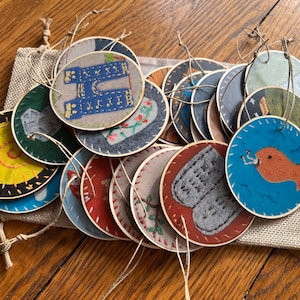 May include: A collection of round, decorative ornaments with various designs, including a fish, clothing, and floral patterns. Each ornament is made of fabric and has a stitched border. They are displayed in a small burlap bag with twine strings for hanging.