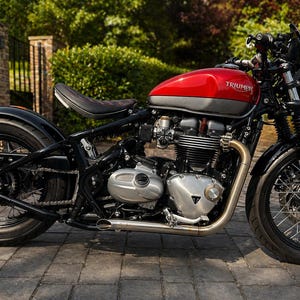 May include: A red and gray Triumph motorcycle with black accents. The motorcycle has a black seat, wheels, and frame. The exhaust pipe is silver. The motorcycle is parked on a brick surface.