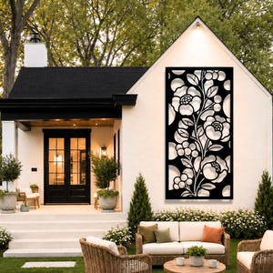 May include: A rectangular black metal wall panel featuring a laser-cut floral pattern with large blossoms, leaves, and small berries. The decorative screen is mounted on the exterior white wall of a house, positioned above a patio seating area.