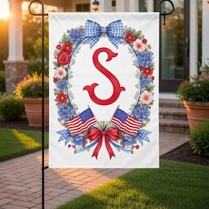 Op de afbeelding: Een witte tuinvaandel met een rode letter "S" omringd door een bloemenkrans met rode, blauwe en roze bloemen. Twee Amerikaanse vlaggen en een rode strik aan de onderkant, met een blauw geruite strik aan de bovenkant.