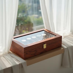May include: A wooden watch display case with a glass top, showcasing several watches. The case is rectangular with a rich, dark brown finish and a gold-colored clasp. It sits on a wooden ledge near a window with sheer white curtains.