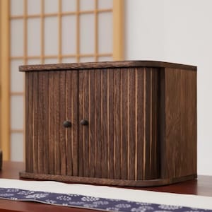 May include: A small, dark brown wooden cabinet with two doors and small, round knobs. The cabinet has a vertical, ridged design on the front and sides. The cabinet is sitting on a table with a patterned runner.