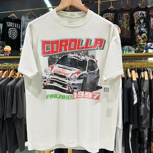 May include: Off-white t-shirt featuring a graphic of a race car with the word "COROLLA" in red and blue. Below the car, the text reads "FINLAND 1997". The shirt is hanging on a wooden hanger.