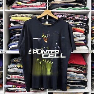 Op de afbeelding: Zwart T-shirt met de tekst "Tom Clancy's Splinter Cell" in het wit. Het shirt heeft een afbeelding van een personage met groen getinte bril. Het T-shirt hangt aan een houten hanger in een kast met opgevouwen kleding.