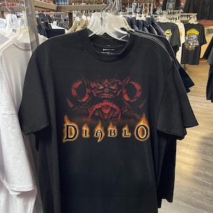 May include: Black t-shirt with a red and orange graphic of a demonic face and the word "Diablo" in a flame-like font. The shirt is displayed on a clothes rack with other garments.