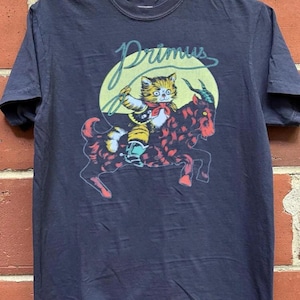 Puede incluir: Camiseta azul marino oscuro con la palabra "Primus" en escritura verde claro. Presenta un gato de dibujos animados montando una cabra. El gato lleva una pajarita roja y sostiene una cuerda. La cabra es roja con manchas negras.