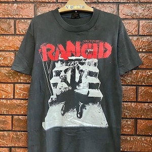 Puede incluir: Camiseta gris oscuro con el nombre de la banda "RANCID" en letras rojas. Debajo, un gráfico en blanco y negro de una figura sobre una estructura. La camiseta está colgada en una percha de madera.