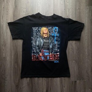 Może przedstawiać: Czarny t-shirt z grafiką osoby w skórzanej kurtce z napisem "RAVEN" w kolorze czerwonym i niebieskim. Koszulka ma wygląd vintage z tekstem w tle.