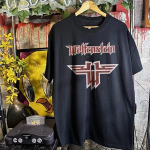 May include: Black t-shirt featuring the word "Wolfenstein" in red and white lettering above a red and white graphic. The shirt is on a wooden hanger. A Nintendo 64 console and a framed Tweety Bird picture are in the background.