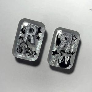 May include: Two rectangular resin pins featuring a silver glitter base, black cat silhouettes, stars, and the letters R and Y. The pins are displayed on a white card with the text x_atelier and small star icons printed in black ink.