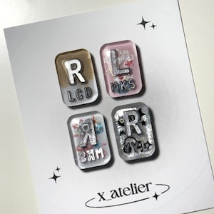 May include: Four rectangular resin charms with rounded corners, each containing silver-coloured letters and small decorative elements. The top left charm features R and LCD, top right L and MKS, bottom left a reversed R and MKS, and bottom right R and VYL.