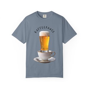 Könnte beinhalten: Blaues T-Shirt mit einem witzigen Design. Ein volles Bierglas steht auf einer weißen Teetasse und Untertasse. Der Text "KAFFEEPAUSE" ist über dem Bierglas angeordnet. Das Shirt ist aus weichem Material.
