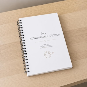 Könnte beinhalten: Ein weißes Notizbuch mit schwarzer Spiralbindung und schwarzem Text auf dem Einband: Unser AUSWANDERUNGSBUCH, gefolgt von einem kleinen Herz, dem Text UNSERE REISE, UNSERE ERINNERUNGEN, UNSER NEUES ZUHAUSE und einer kleinen Strichzeichnung eines Globus.
