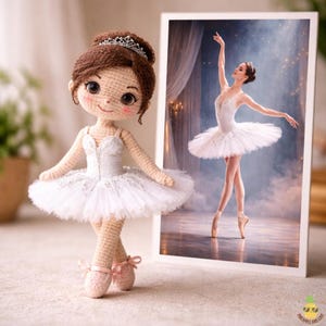 May include: A crocheted ballerina doll with a white tutu, tiara, and pink ballet shoes stands next to a framed photo of a ballerina. The doll has brown hair and rosy cheeks. The photo shows a ballerina in a white tutu.