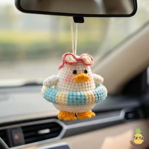 May include: A handmade crochet duck car charm. The duck is white with pink cheeks, a yellow beak, and small black eyes. It wears pink-rimmed sunglasses and a blue and yellow striped inner tube. The duck has yellow feet.