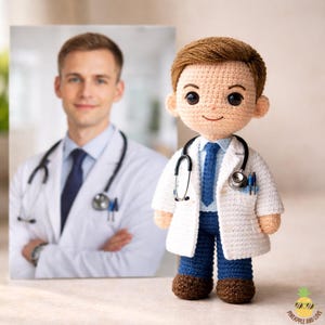Könnte beinhalten: Gehäkelte Arztpuppe mit weißem Kittel, blauer Krawatte und Stethoskop. Die Puppe hat braunes Haar und steht neben einem gerahmten Foto eines Arztes. Ein handgefertigter Artikel.