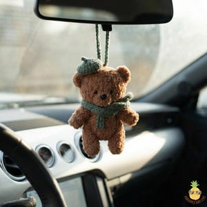 May include: A brown teddy bear car charm with a green beret and scarf. The plush bear is hanging from the rearview mirror. The car's dashboard and steering wheel are visible in the background.