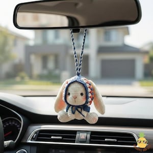 May include: A handmade crochet bunny car charm. The bunny is white with a blue and orange bonnet and a blue bow. The charm hangs from a rearview mirror in a car.