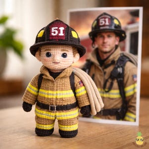 May include: A crocheted firefighter doll wearing a brown uniform with yellow stripes, a black helmet with the number 51, and a black belt. The doll has a tan hose over its shoulder and a smiling face. A photo of a firefighter is in the background.