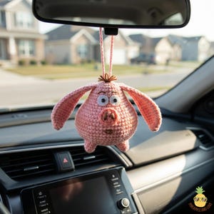 Puede incluir: Un adorno de coche de cerdo rosa tejido a ganchillo, colgado del espejo retrovisor de un coche. El cerdo tiene orejas grandes, ojos grandes y un hocico detallado. El adorno está suspendido por una cuerda rosa, añadiendo un toque de fantasía al interior del coche.