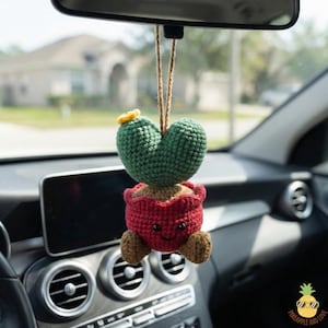 May include: A handmade, crocheted cactus car charm. The charm features a green heart-shaped cactus with a yellow flower, sitting in a red pot with a smiling face. The charm hangs from a brown string, perfect for adding a touch of whimsy to any vehicle.
