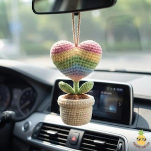 May include: A handmade crochet car charm featuring a rainbow heart-shaped flower with green leaves and a beige pot. The charm hangs from a rearview mirror, adding a touch of color and whimsy to the car's interior.