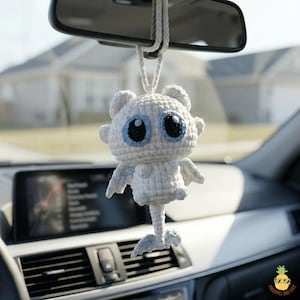 May include: A white crocheted car charm shaped like a cartoon dragon, hanging from a car's rearview mirror. The dragon has large blue and black eyes, small wings, and a tail. The charm is suspended by a white braided cord.