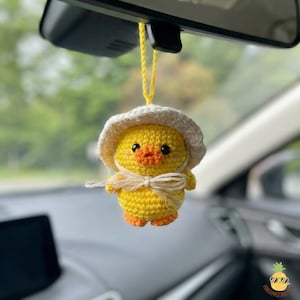 May include: A handmade yellow crochet duck wearing a white hat and a bow. The duck has orange feet and a beak. It is hanging from a yellow string, likely as a car charm.