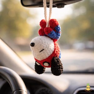 May include: A handmade crochet car charm in the shape of a cartoon character, hanging from a white string. The charm features a white head, red body, blue accents, and black details. The charm is hanging from a car's rearview mirror.