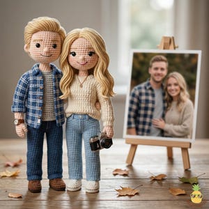 Puede incluir: Un par de muñecos de ganchillo: un hombre con camisa de cuadros azul y vaqueros, y una mujer con jersey crema y vaqueros, sosteniendo una pequeña cámara. Una foto enmarcada de una pareja está detrás de ellos. Los muñecos están sobre una superficie de madera.