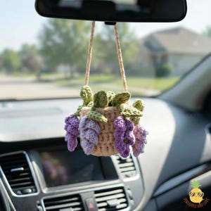 May include: A handmade crochet car charm featuring a beige basket filled with purple grape clusters and green leaves. The charm hangs from a rearview mirror, adding a touch of nature-inspired decor to the car's interior.