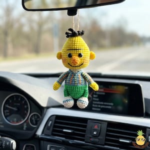 May include: A handmade yellow crochet doll with black hair, a smiling face, and a blue, green, and orange outfit. The doll is hanging from a string, likely as a car accessory. The background is blurred, suggesting it's inside a vehicle.