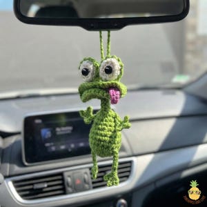 May include: A green crocheted frog car charm with large, googly eyes and a pink tongue hanging out. The frog is suspended from a car's rearview mirror. The charm is handmade and adds a whimsical touch to the vehicle's interior.