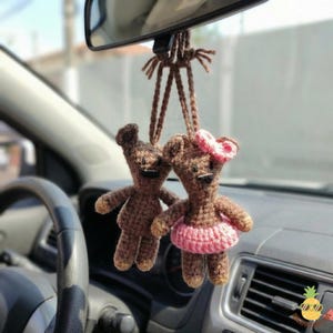 Può includere: Due ciondoli per auto a forma di orsetto all'uncinetto marrone appesi allo specchietto retrovisore di un'auto. Un orsetto ha un fiocco rosa e una gonna rosa. L'altro orsetto è semplice. Gli orsetti sono appesi davanti al cruscotto e al volante di un'auto.
