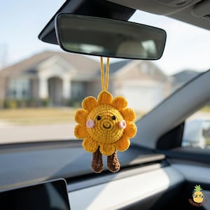 Op de afbeelding: Een gele gehaakte zon-auto-hanger met een lachend gezicht, roze wangen en bruine benen. De zon-hanger hangt aan een gele draad voor de achteruitkijkspiegel van een auto. De hanger is handgemaakt.