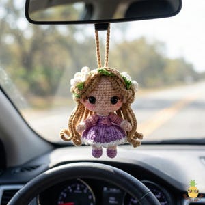 May include: A crocheted doll car charm with long, curly brown hair, large black eyes, and a purple dress. The doll is adorned with a floral crown and hangs from a beige string, suspended from a car's rearview mirror.