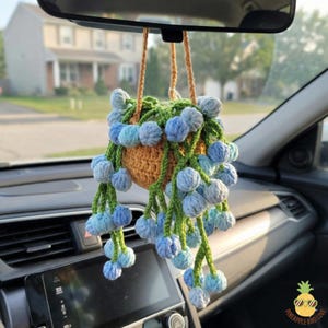 May include: A handmade crochet hanging car plant with blue and green accents. The plant is suspended from the rearview mirror by a tan cord. The crochet plant pot is tan and filled with green leaves and blue pom-poms.