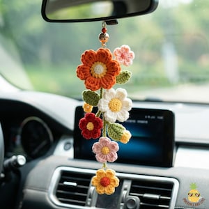 May include: A handmade crochet car charm featuring a cascade of colorful flowers. The charm includes orange, white, red, pink, and yellow flowers with green leaves, hanging from a rearview mirror.