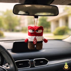May include: A crocheted car charm featuring a red character with an angry expression. The charm is wearing a white shirt, brown pants, and a red tie. It hangs from a string attached to a car's rearview mirror.