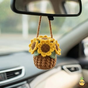 Könnte beinhalten: Ein handgefertigter gehäkelter Autoanhänger mit einem Strauß Sonnenblumen. Die Sonnenblumen haben gelbe Blütenblätter und braune Zentren mit grünen Blättern. Die Blumen befinden sich in einem braunen Korb, der an einer braunen Schnur hängt.