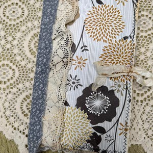 May include: A rectangular journal featuring a cover with a white background, vertical gray lines, and a floral pattern of mustard yellow and dark brown dahlia-like flowers. The spine is accented with blue floral fabric and cream lace, finished with a cream ribbon tie.