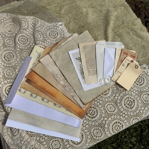 May include: A collection of assorted paper ephemera including tea-stained sheets, vintage-style book pages, white envelopes, and cream-colored shipping tags with string. The papers are layered in a fan shape, featuring various textures and shades of beige.