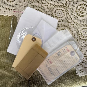 May include: A collection of stationery items including a white envelope, a light tan shipping tag with a string, a small brown paper envelope, and a translucent glassine envelope containing a printed tag with the text No. 572832 and lines for details.