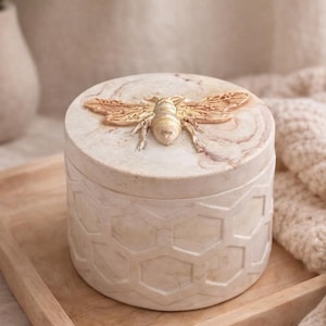 May include: A round, beige decorative box with a lid featuring a gold-coloured bee. The box has a honeycomb pattern on the sides and sits on a wooden tray. The lid has a marble-like design.