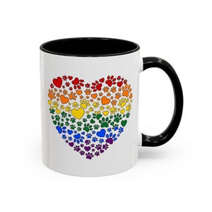May include: White ceramic mug with a black interior and handle. The mug features a heart design composed of colorful paw prints and hearts in a rainbow spectrum. A cheerful and vibrant design.