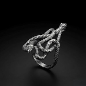 Peut inclure: Une bague en argent foncé présentant un motif détaillé de deux serpents entrelacés. La surface métallique présente un aspect texturé et écailleux sur tout le corps des serpents, leurs têtes étant positionnées au sommet de la structure.
