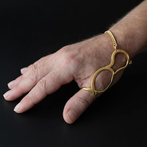 May include: A gold-colored hand accessory featuring a ring for the thumb and a bracelet chain. The design includes an open, abstract shape that extends across the back of the hand. The accessory is made of a shiny, reflective metal.