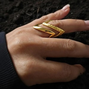May include: A gold-coloured ring featuring a double-band design with a V-shaped silhouette. One band has a smooth, polished finish, while the other band displays a textured, hammered surface. The ring is worn on a finger, showcasing its geometric shape.