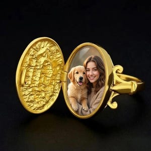 Könnte beinhalten: Ein goldener Medaillonring mit einem ovalen Klappfach. Im Inneren befindet sich ein Foto einer Person mit langem braunem Haar, die lächelt und einen goldenen Welpen hält. Die Außenseite des Medaillondeckels weist eine texturierte, unebene Oberfläche auf.