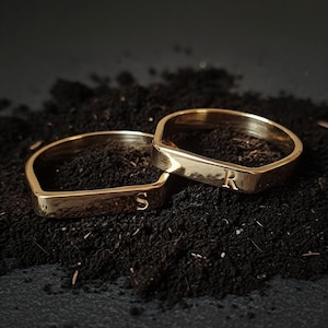 Puede incluir: Dos anillos dorados, cada uno con una superficie rectangular plana, descansan sobre una superficie oscura y texturizada. Un anillo muestra la letra "S", mientras que el otro muestra la letra "R". Los anillos tienen un acabado pulido.